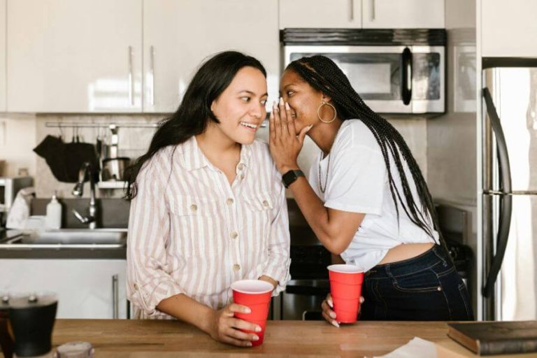 Dos mujeres conversando y compartiendo el chisme en una cocina, susurrando con complicidad.