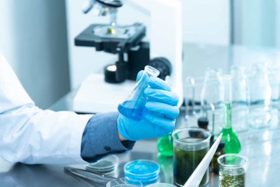 Investigador con guantes azules sosteniendo un matraz en un laboratorio con microscopio y soluciones químicas, representando la evidencia científica en el debate sobre pseudoterapias