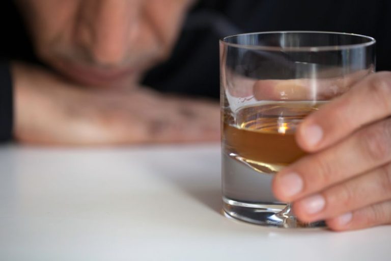 Mano de un hombre sosteniendo un vaso de alcohol, con el rostro desenfocado, simbolizando el consumo y la pérdida de control.
