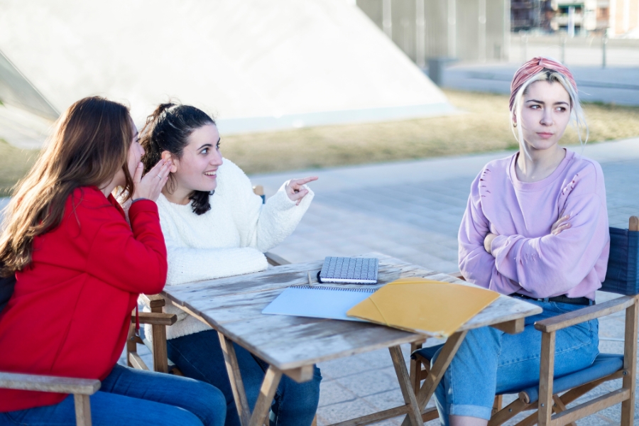 Dos adolescentes critican a una compañera que permanece con los brazos cruzados y la mirada apartada, reflejando el impacto emocional de la crítica durante la adolescencia.