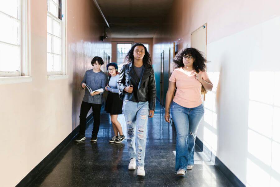 Adolescentes caminando por un pasillo escolar, dos al fondo observando, escena de bullying.