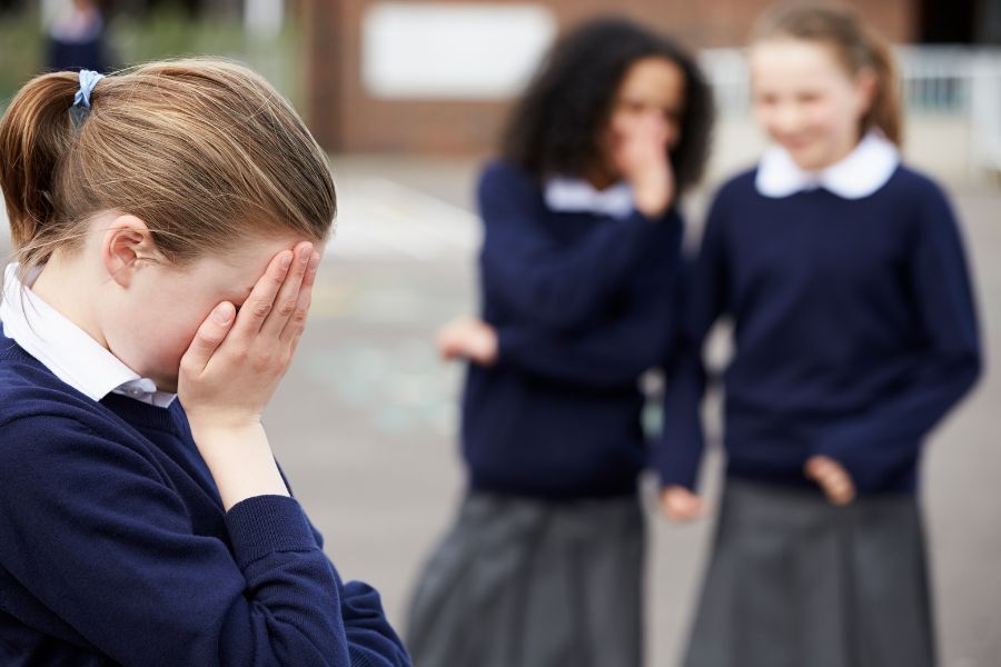 Niña en uniforme escolar llorando por acoso escolar mientras dos compañeras se burlan de ella en el patio del colegio.