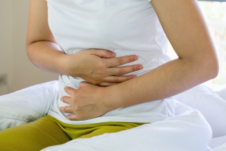 Persona sentada en una cama, con ambas manos presionando firmemente su abdomen bajo, mostrando signos de dolor o malestar estomacal.